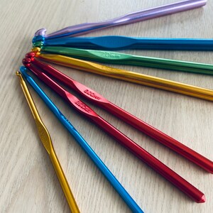 May include: A set of colorful crochet hooks, including purple, blue, green, gold, and red. The hooks are arranged in a fan shape on a wooden surface. The hooks are labeled with their sizes in millimeters.