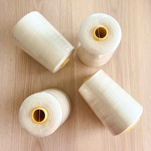 May include: Four spools of off-white thread are arranged on a light wood surface. The thread is wrapped in clear plastic. The spools have yellow centres. The thread is likely for sewing or crafting.