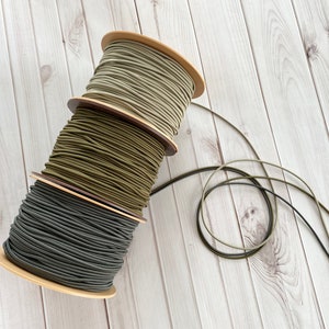 May include: Three spools of elastic cord in different shades of green, wound around cardboard spools. The spools are stacked on top of each other. The elastic cord is a popular crafting material.