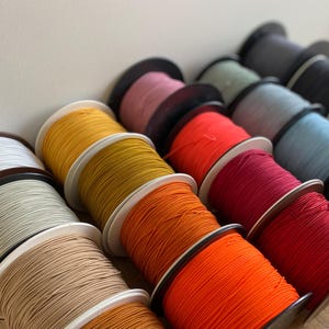 Può includere: Una collezione di bobine di cordoncino elastico colorato, tra cui bianco, beige, giallo, arancione, rosso e blu. Le bobine sono disposte in fila, con i colori che passano gradualmente da chiaro a scuro.