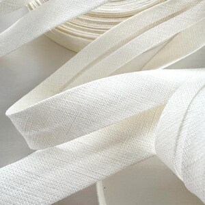 May include: Close-up of white bias tape, with a textured weave, in a roll and unraveled. The tape is a soft, off-white colour, and the image highlights the fabric's texture and the tape's flexibility. Ideal for sewing and crafting projects.