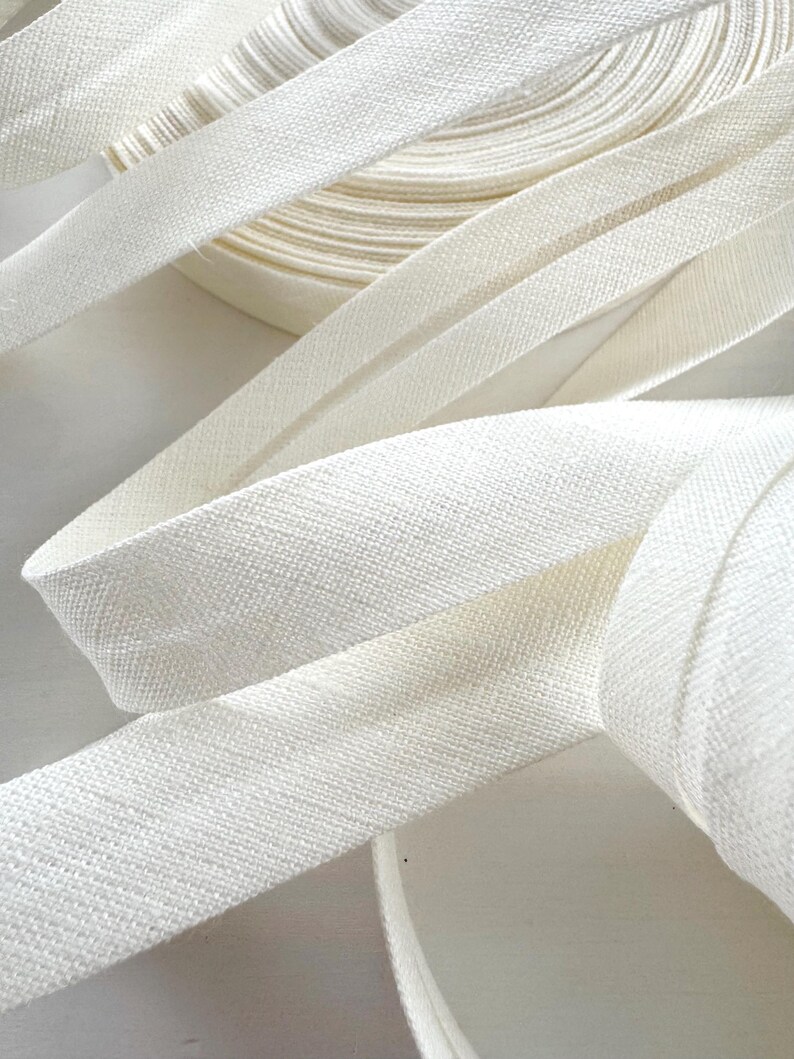 May include: Close-up of white bias tape, with a textured weave, in a roll and unraveled. The tape is a soft, off-white colour, and the image highlights the fabric's texture and the tape's flexibility. Ideal for sewing and crafting projects.