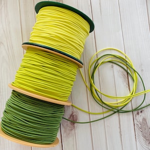 Könnte beinhalten: Drei Spulen mit elastischem Band, eine grün und zwei gelb, sind auf einer Holzoberfläche gestapelt. Die Spulen sind mit elastischem Band umwickelt.
