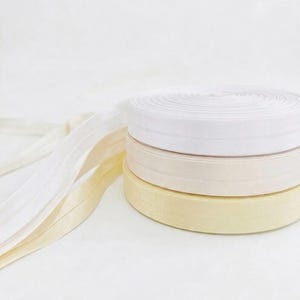Pode incluir: Três rolos empilhados de fita elástica plana em tons de branco, creme e amarelo pálido. Cada rolo apresenta uma textura tecida lisa com uma linha de vinco central que corre horizontalmente ao longo do comprimento das pontas estendidas.