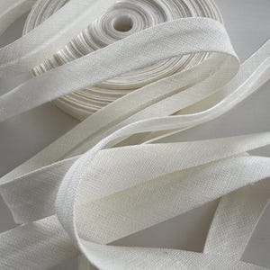 May include: A roll of white bias binding with a woven texture. The tape is unwound in several places, revealing the fabric's detail. The binding is likely used for sewing, crafting, or other fabric projects.