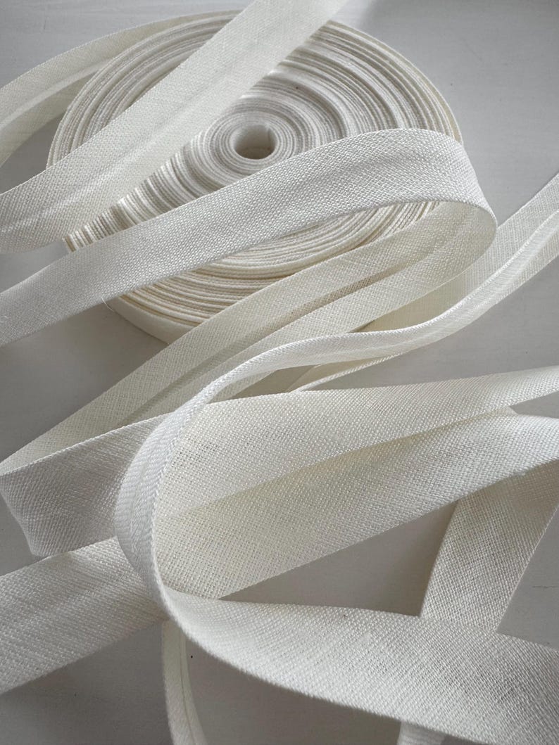 May include: A roll of white bias binding with a woven texture. The tape is unwound in several places, revealing the fabric's detail. The binding is likely used for sewing, crafting, or other fabric projects.