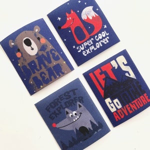 Op de afbeelding: Vier blauwe stoffen patches met kleurrijke illustraties en tekst. De patches tonen een beer, een vos, een wolf en een huis. De tekst op de patches luidt "Brave Bear", "Super Cool Explorer", "Let's Go On An Adventure" en "Forest Explorer".