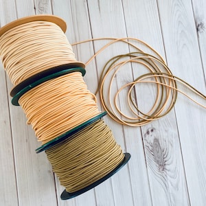 Pode incluir: Três rolos de corda, um bege, um bege claro e um verde oliva, estão empilhados em uma superfície de madeira. As cordas são enroladas em torno de rolos com etiquetas verdes e brancas.