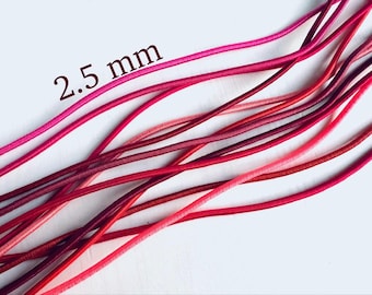 May include: Close-up of several strands of 2.5 mm diameter elastic cord in various shades of red and pink. The cords are arranged diagonally across a white background, creating a visually appealing display. The text "2.5 mm" is visible in the upper left corner.