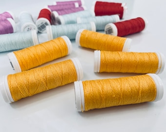 Könnte beinhalten: Eine Sammlung von Garnrollen in verschiedenen Farben, darunter Gelb, Rot, Blau und Lila. Die Rollen sind auf einer weißen Oberfläche angeordnet.