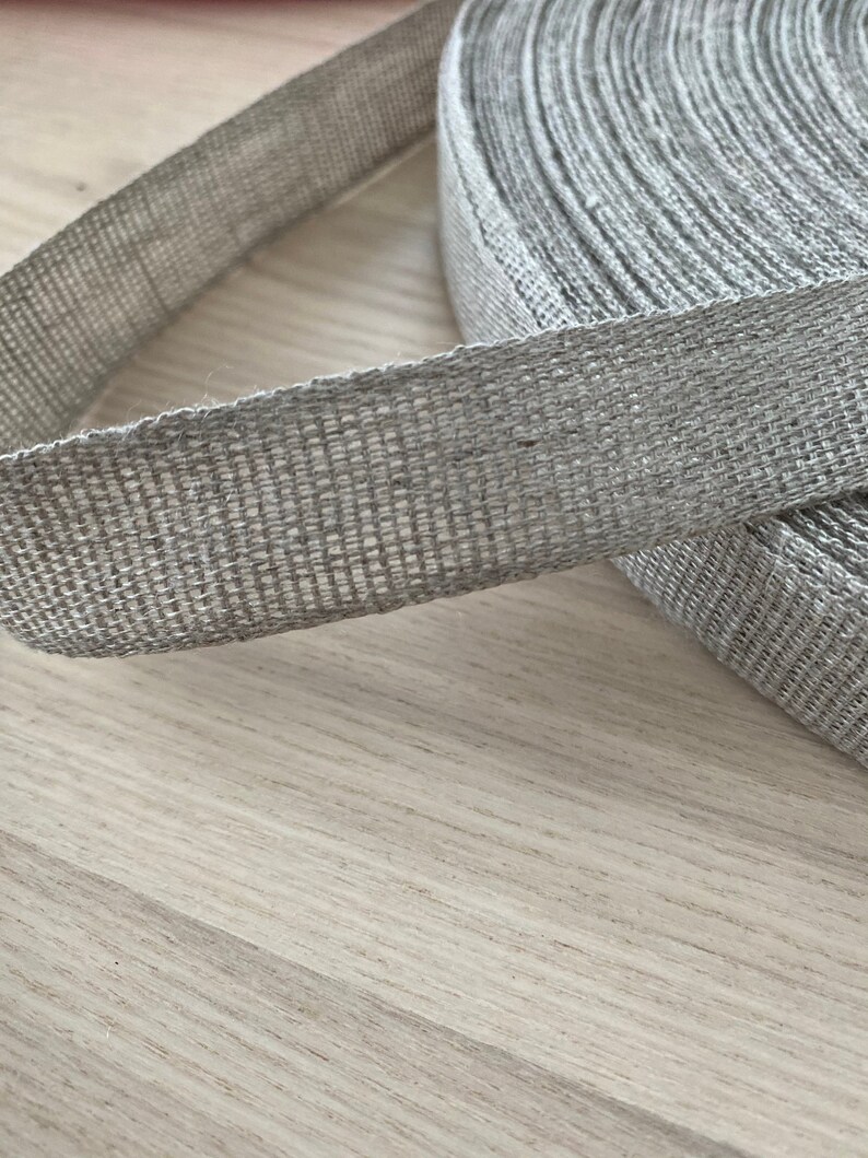 May include: A roll of natural linen ribbon, woven with a textured pattern. The ribbon is light gray and has a natural, rustic look.