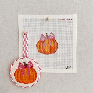 May include: A needlepoint canvas featuring an orange pumpkin with a pink bow, accompanied by a circular ornament displaying the same pumpkin design with a pink and white twisted cord hanger. The canvas includes a color palette guide at the top.