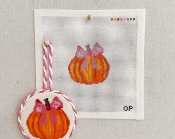 Puede incluir: Un lienzo de punto de aguja con una calabaza naranja con un lazo rosa, acompañado de un adorno circular que muestra el mismo diseño de calabaza con un cordón trenzado rosa y blanco. El lienzo incluye una guía de colores en la parte superior.