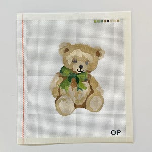 May include: A square needlepoint canvas featuring a tan teddy bear with a green bow around its neck. The bear is centered on a white grid background. A vertical red line runs along the left edge, and the letters OP are printed in the bottom right corner.