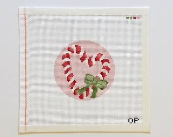 Puede incluir: Un lienzo cuadrado de punto de aguja con un diseño de bastón de caramelo en forma de corazón centrado sobre un fondo circular rosa pálido. El bastón tiene rayas rojas y blancas y un lazo verde. Las letras OP están bordadas en la esquina inferior.