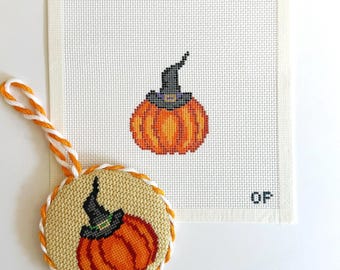 Puede incluir: Un lienzo cuadrado de punto de aguja con una calabaza naranja bordada en punto de cruz con un sombrero de bruja negro, junto a un adorno circular terminado con el mismo diseño, bordeado por un cordón trenzado naranja y blanco con lazo.