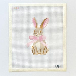 May include: Needlepoint canvas featuring a seated bunny with a pink bow. The bunny is tan and brown, with pink-lined ears. The background is off-white, with the letters "OF" in the bottom right corner.