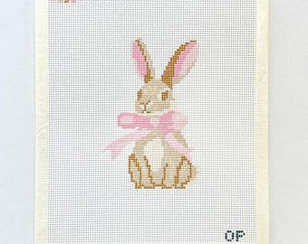 May include: Needlepoint canvas featuring a seated bunny with a pink bow. The bunny is tan and brown, with pink-lined ears. The background is off-white, with the letters "OF" in the bottom right corner.