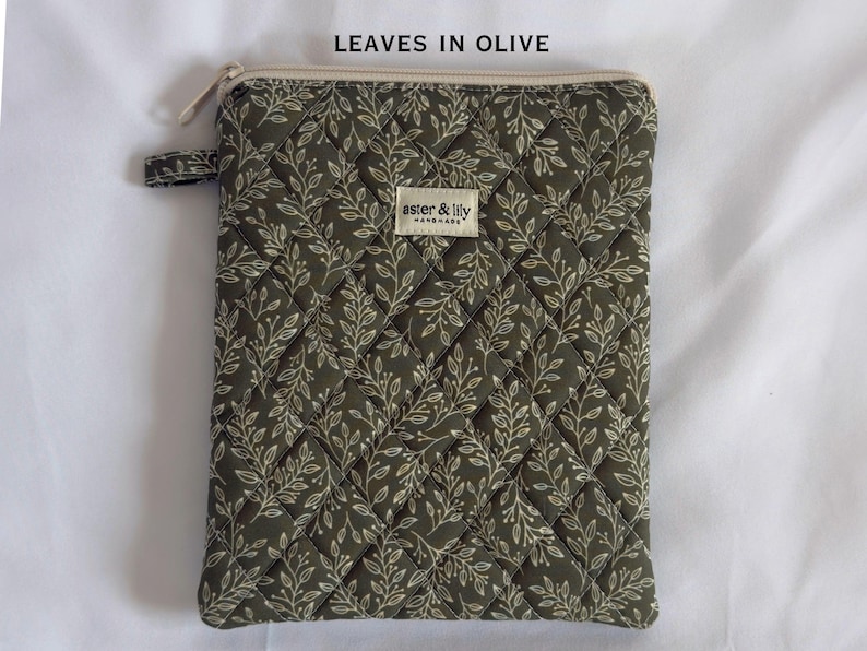 May include: A rectangular olive green zipped pouch featuring a quilted diamond pattern and a white leaf print. A small fabric tag labelled aster & lily is centred on the front. The text LEAVES IN OLIVE is printed above the pouch.