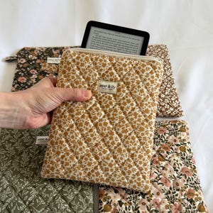 May include: A quilted fabric sleeve with a mustard yellow floral pattern and a white zip closure. A small rectangular label reads aster & lily. The sleeve contains a black e-reader displaying text. Other quilted sleeves with floral patterns are visible.