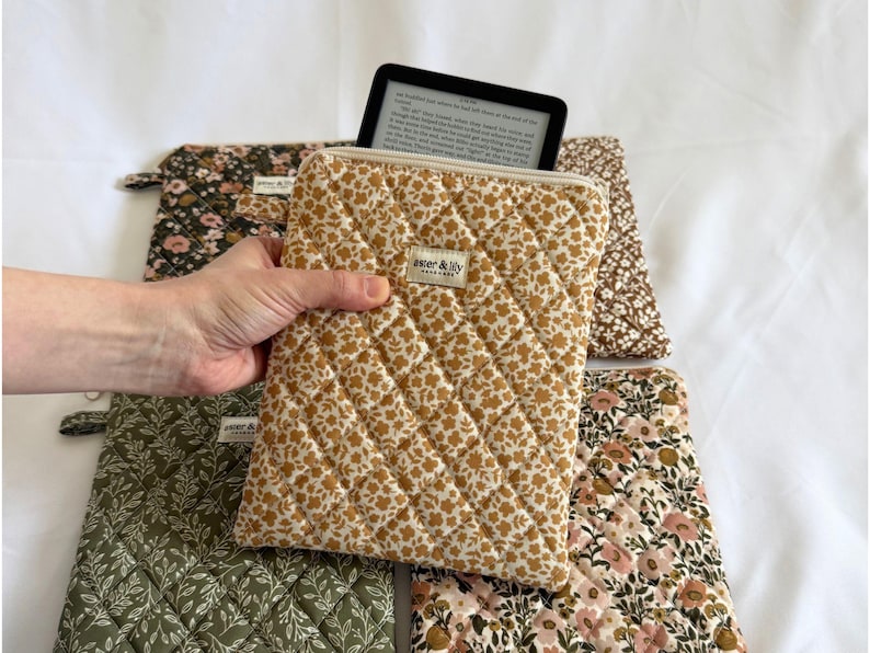May include: A quilted fabric sleeve with a mustard yellow floral pattern and a white zip closure. A small rectangular label reads aster & lily. The sleeve contains a black e-reader displaying text. Other quilted sleeves with floral patterns are visible.