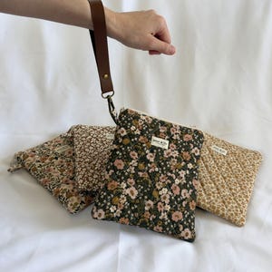 May include: Four quilted fabric pouches with floral patterns in shades of green, brown, and cream. Each pouch features a zip closure, a small rectangular label reading "aster & lily", and one pouch is attached to a brown leather wrist strap.