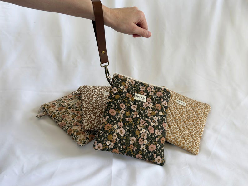 May include: Four quilted fabric pouches with floral patterns in shades of green, brown, and cream. Each pouch features a zip closure, a small rectangular label reading "aster & lily", and one pouch is attached to a brown leather wrist strap.