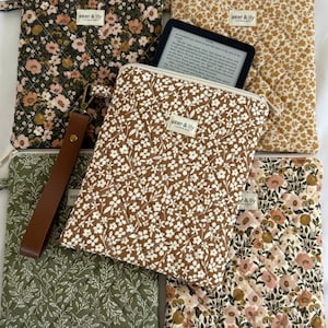 May include: Five quilted fabric pouches with zip closures and wrist straps, featuring various floral patterns in olive, mustard, brown, and cream. Each pouch has a small rectangular label reading aster & lily handmade. One pouch holds an e-reader.
