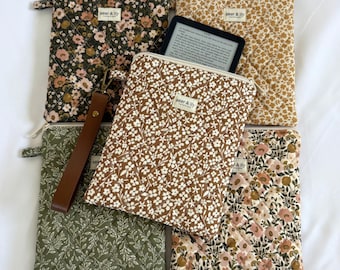 May include: Five quilted fabric pouches with zipper closures and wrist straps, featuring various floral patterns in olive, mustard, brown, and cream. Each pouch has a small rectangular label reading aster & lily handmade. One pouch holds an e-reader.