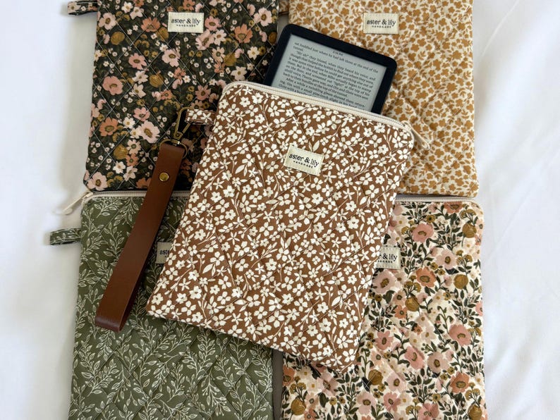 May include: A collection of five quilted fabric sleeves for e-readers, each featuring floral patterns in shades of olive, mustard, and brown. One sleeve holds a black e-reader, and each has a small label reading aster & lily. A brown wrist strap is attached.