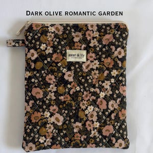 May include: A rectangular quilted pouch in dark olive with a floral pattern in shades of pink, cream, and brown. It features a top zip closure, a small side tab, and a central cream-coloured label with the text "aster & lily HANDMADE".