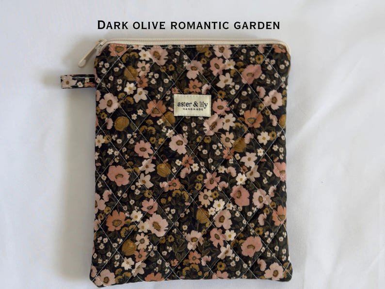 May include: A rectangular quilted pouch in dark olive with a floral pattern in shades of pink, cream, and brown. It features a top zip closure, a small side tab, and a central cream-coloured label with the text "aster & lily HANDMADE".