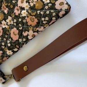 May include: A quilted pouch featuring a black fabric base with a floral pattern in shades of pink, white, and mustard yellow. The pouch includes a detachable brown wrist strap secured with a brass-toned metal rivet and a matching lobster clasp.