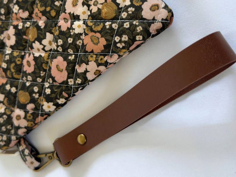 May include: A quilted pouch featuring a black fabric base with a floral pattern in shades of pink, white, and mustard yellow. The pouch includes a detachable brown wrist strap secured with a brass-toned metal rivet and a matching lobster clasp.