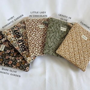 May include: Four quilted pouches with zip closures and small fabric labels reading 'pear & lily'. Patterns include dark olive floral, ivory floral marked 'SOLD OUT', chocolate brown floral, olive green leaves, and tan daisy print.