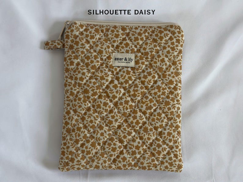 May include: A rectangular quilted pouch featuring a mustard yellow and white floral pattern. The top includes a zip closure and a small fabric tab. A rectangular label in the centre reads aster & lily HANDMADE. Text above reads SILHOUETTE DAISY.