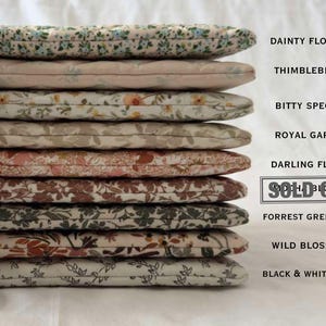May include: A stack of nine quilted fabric pouches with various floral and vine patterns. Text labels include Dainty Flowers, Thimbleberry, Bitty Specter, Royal Garden, Darling Floral, Sold Out, Forrest Green Blooms, Wild Blossoms, and Black & White Vines.