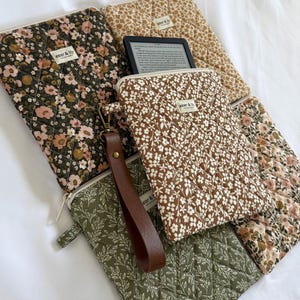 May include: A collection of quilted fabric pouches featuring floral and leaf patterns in shades of olive green, brown, and cream. Each pouch has a zip closure and a small label reading "aster & lily". One pouch includes a brown leather wrist strap.