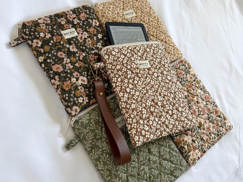 May include: A collection of quilted fabric pouches featuring floral and leaf patterns in shades of olive green, brown, and cream. Each pouch has a zip closure and a small label reading "aster & lily". One pouch includes a brown leather wrist strap.