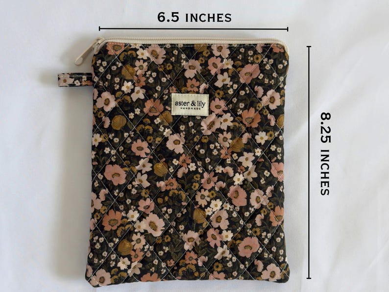 May include: A quilted pouch with a dark floral pattern in shades of pink, brown, and cream. It features a zip closure, a small side tab, and a label reading aster & lily. Dimensions are 16.51 cm wide by 20.96 cm high.