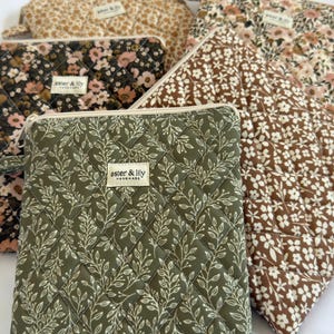 May include: A collection of five quilted pouches featuring various floral and leaf patterns in olive green, brown, cream, and black. Each rectangular pouch has a zip closure and a small fabric label reading aster & lily handmade.