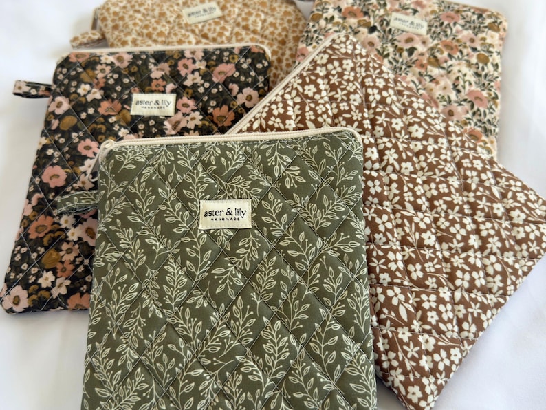 May include: A collection of five quilted pouches featuring various floral and leaf patterns in olive green, brown, cream, and black. Each rectangular pouch has a zip closure and a small fabric label reading aster & lily handmade.
