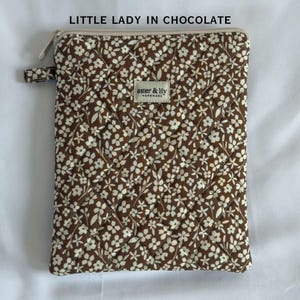 May include: A rectangular quilted pouch in a chocolate brown fabric featuring a white floral pattern. The top features a zip closure and a small fabric label that reads aster & lily handmade. The text LITTLE LADY IN CHOCOLATE is visible.