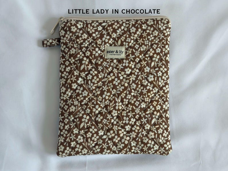 May include: A rectangular quilted pouch in a chocolate brown fabric featuring a white floral pattern. The top features a zip closure and a small fabric label that reads aster & lily handmade. The text LITTLE LADY IN CHOCOLATE is visible.