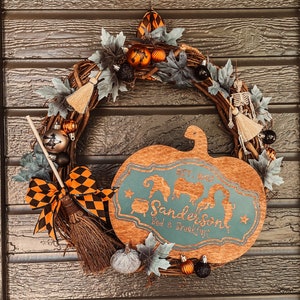 Könnte beinhalten: Ein Halloween-Kranz mit einem hölzernen Kürbis-Schild, auf dem "Get Spooky Sanderson's Bed & Breakfast" steht. Der Kranz ist mit Herbstlaub, orangefarbenen und schwarzen Ornamenten und einem kleinen Besen verziert.