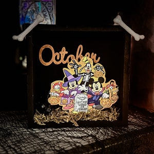 May include: A black wooden box with a gold inscription that reads "October". The box features a Halloween-themed image of Mickey Mouse, Minnie Mouse, Goofy, and Donald Duck dressed in costumes. The image is surrounded by faux moss and bone-shaped decorations.