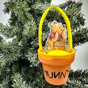 Könnte beinhalten: Ein Winnie Puuh Weihnachtsornament mit einem gelben Topf, auf dem das Wort "Hunnny" steht. Winnie Puuh hält einen Honigtopf und sitzt auf dem Rand des Topfes.