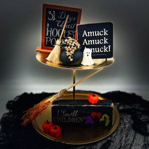 Könnte beinhalten: Ein zweistöckiges goldenes Serviertablett mit Halloween-Dekorationen. Die obere Etage enthält einen Kessel, einen Besen und ein Schild mit der Aufschrift "Amuck Amuck Amuck!". Die untere Etage hat ein Schild mit der Aufschrift "I Smell Children" und kleine Kürbisse.