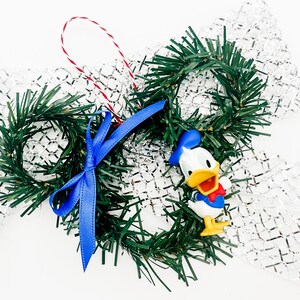 Puede incluir: Un adorno navideño con una pequeña figura de plástico de Donald Duck con un sombrero de marinero azul y sosteniendo un lazo rojo. El adorno está rodeado de una corona verde con una cinta azul.
