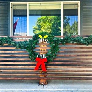 May include: A wooden porch railing decorated with a green garland and a wreath with a red bow and a monster face cutout. The monster face has yellow eyes and white teeth.